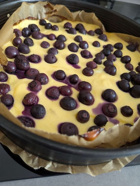 Frisch gebackener Blaubeerkuchen in einer runden Springform mit leicht gebräuntem Quarkguss und ganzen Blaubeeren, ausgelegt mit Backpapier auf einem Herd. 