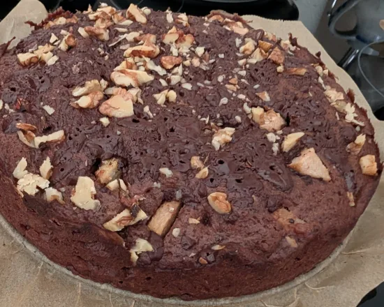 Ganzer Schoko-Birnenkuchen auf Backpapier, obenauf gehackte Walnüsse, Birnenstücke und geschmolzene Schokolade