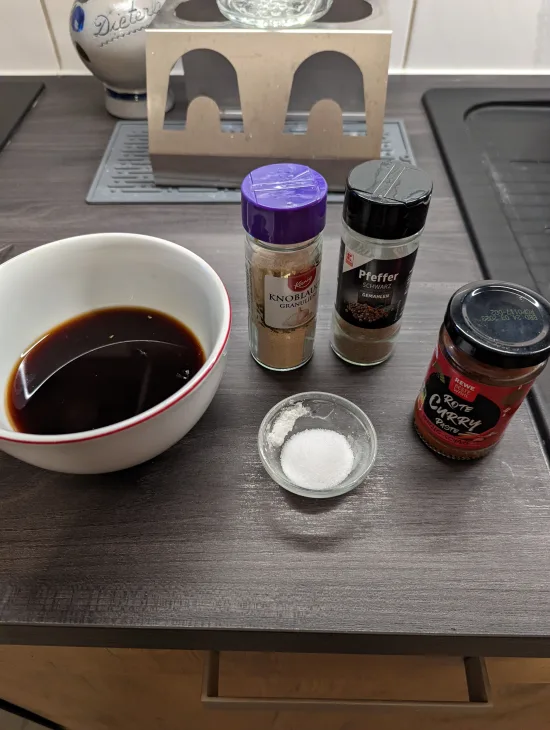 Zutaten der Sauce: Schale mit Sojasauce, Orangen- und Zitronensaft, daneben granulierter Knoblauch, schwarzer Pfeffer, Zucker und rote Currypaste