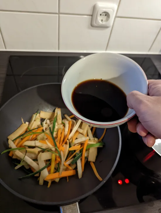 Die dunkle Sauce wird aus einer Schale zum angebratenen Gemüse in den Wok gegossen