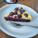 Ein Stück selbstgebackener Blaubeerkuchen mit dunklem Schokoboden, cremiger Quarkschicht und frischen Blaubeeren obendrauf auf einem hellen Teller (daneben eine Gabel); im Hintergrund steht unscharf ein weißer Becher mit der Aufschrift „Cabo Verde“.