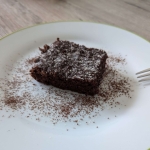 Ein saftiges Stück schokoladiger Zucchinikuchen auf einem weißen Teller, leicht mit Puderzucker und Kakaopulver bestäubt.